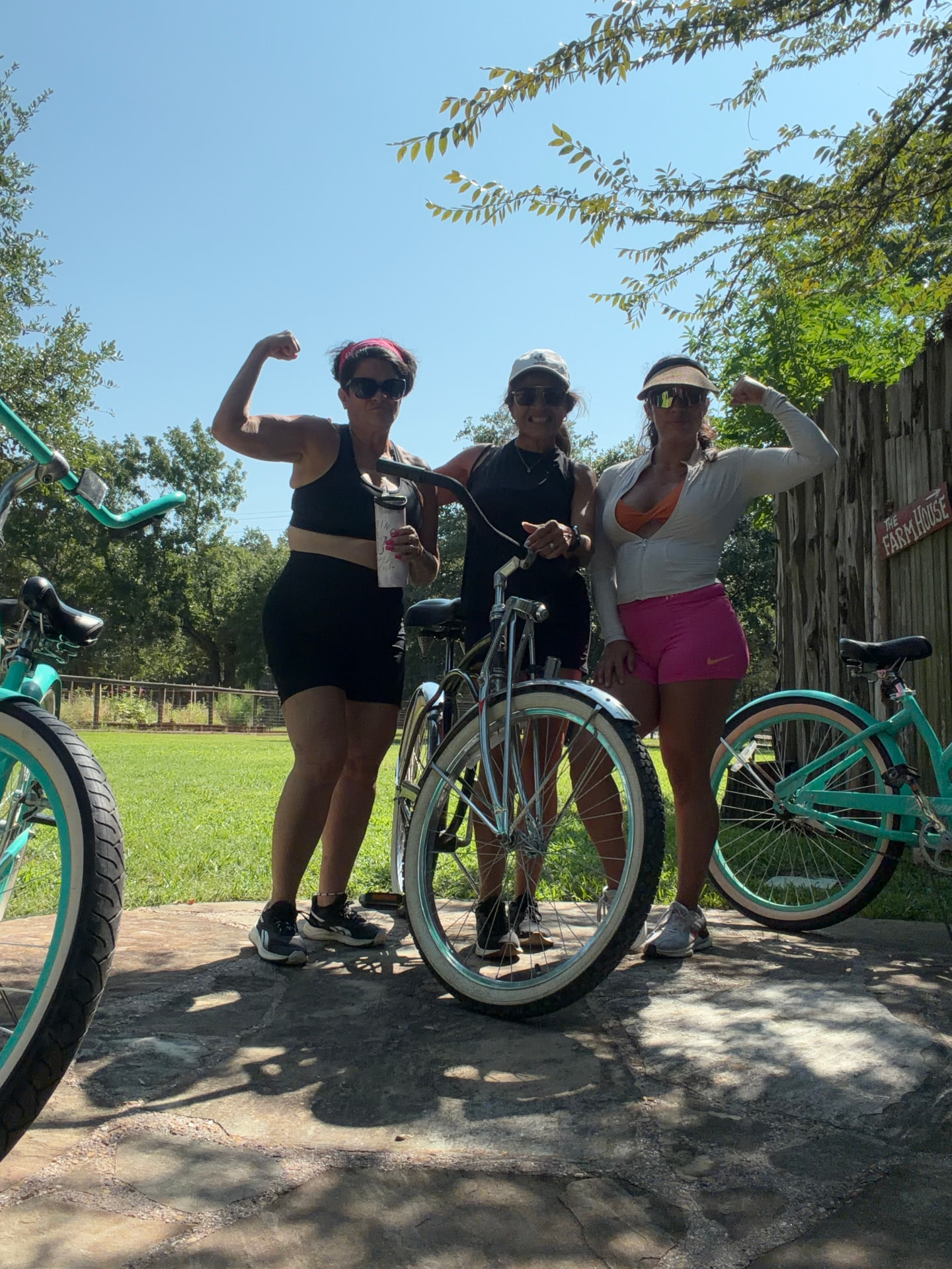 Three women flexing proudly beside teal cruiser bikes on a sunny stone path